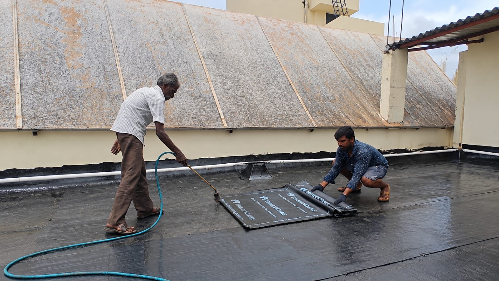 Two applicators heat-fusing a bitumen membrane roll with a gas torch on a rooftop