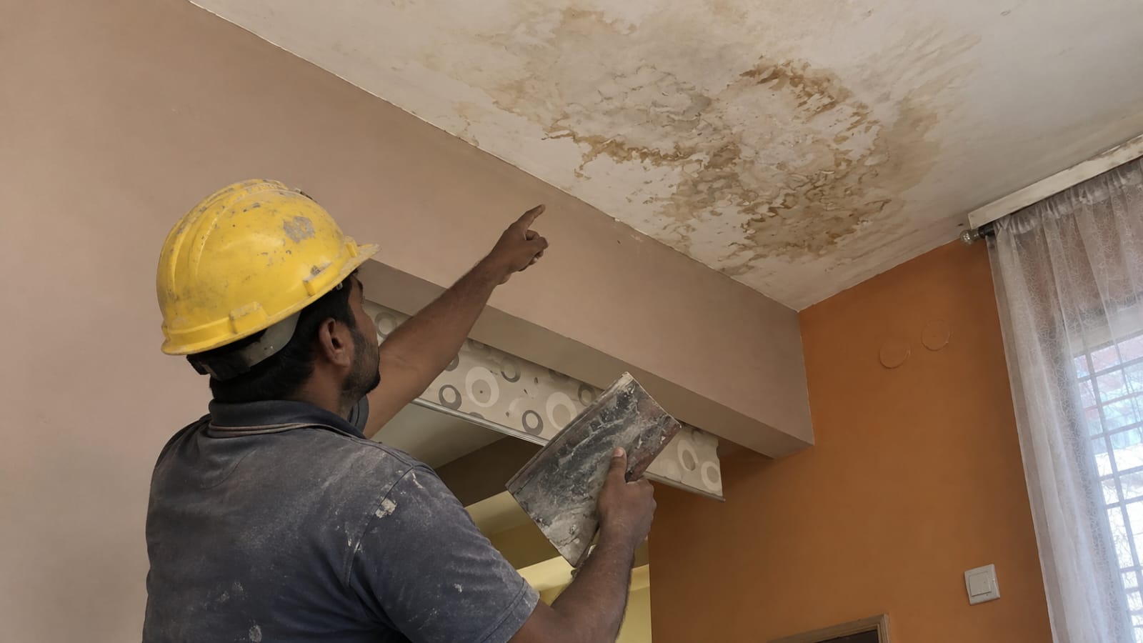 Worker examining a brown water-stained damp patch spreading across an interior ceiling