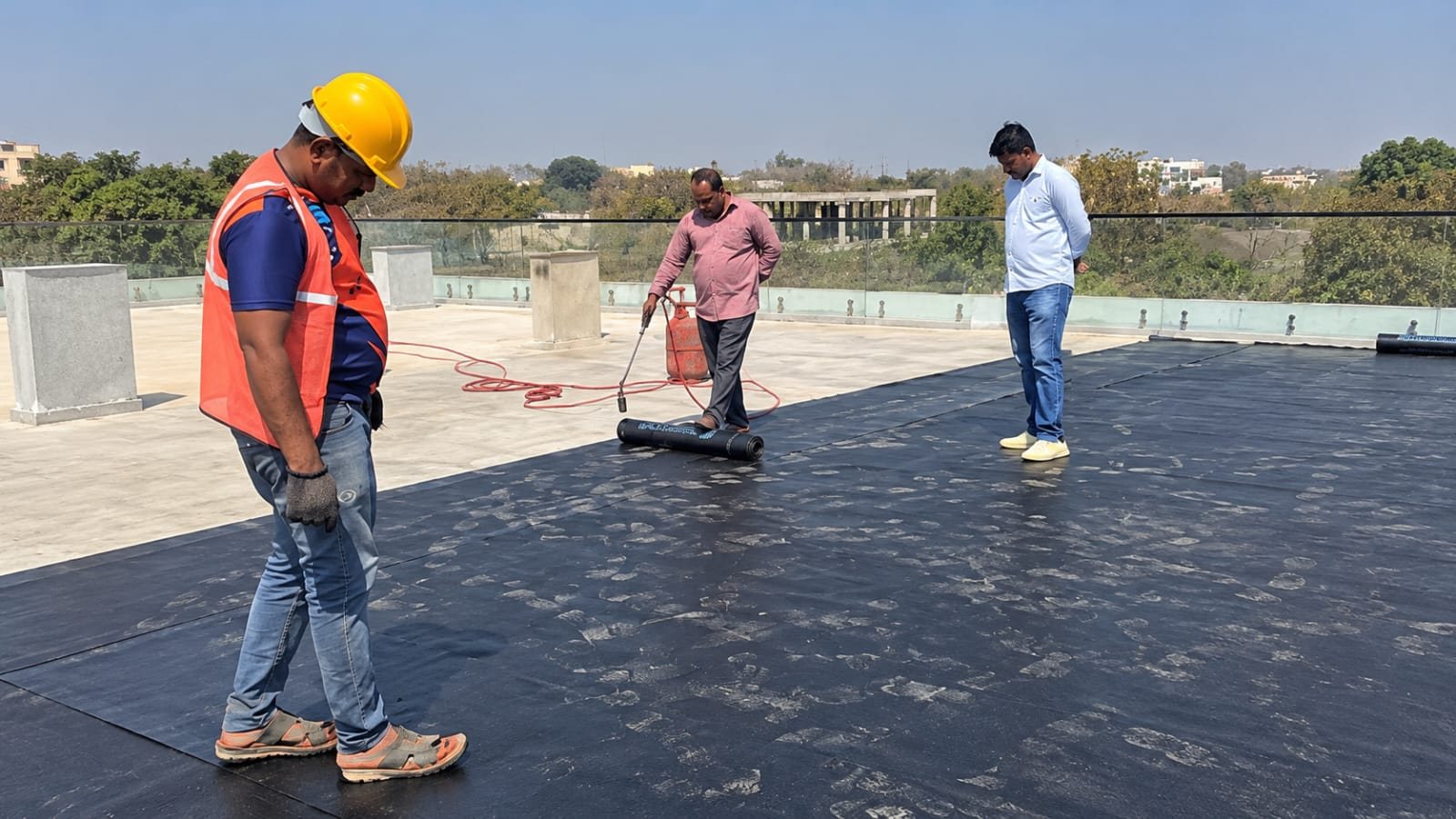 Three workers on a freshly coated black rooftop with a glass parapet railing and trees behind